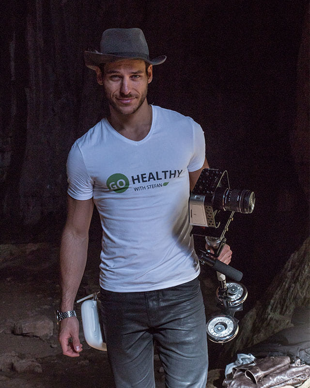 A man wearing a gray fedora hat and a white t-shirt with a "GO HEALTHY WITH STEFAN" logo smiles while holding a professional camera mounted on a stabilizer rig in a dimly lit, cave-like environment.