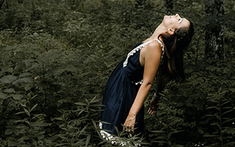 A woman in a dark dress stands among dense green foliage, her head tilted back, looking upward.